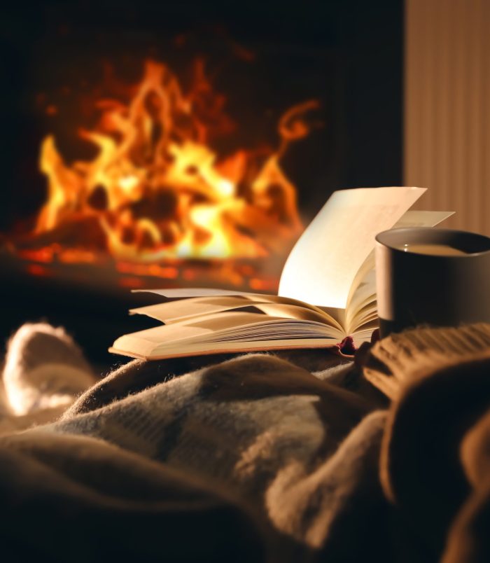 Woman,With,Cup,Of,Drink,And,Book,Near,Fireplace,At